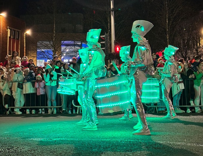 Performers in silver costumes with green-lit drums playing in front of cheering crowd.