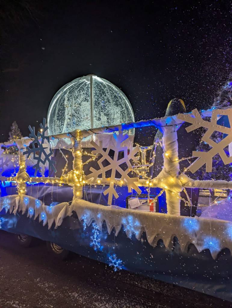 Parade float with illuminated snow globe, snowflake lights, and wintry decorations.