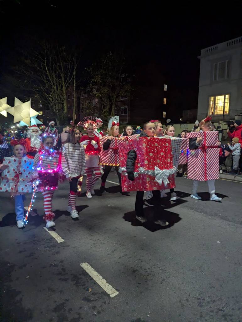 Children and adults in illuminated gift box costumes walking in Christmas parade.