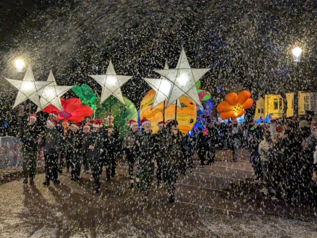 Parade participants carrying glowing star lanterns through snow-covered street with inflatable decorations.