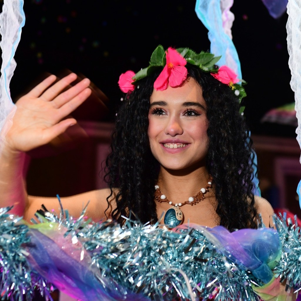 Parade performer in floral crown and tropical outfit with tinsel and beads, smiling and waving.
