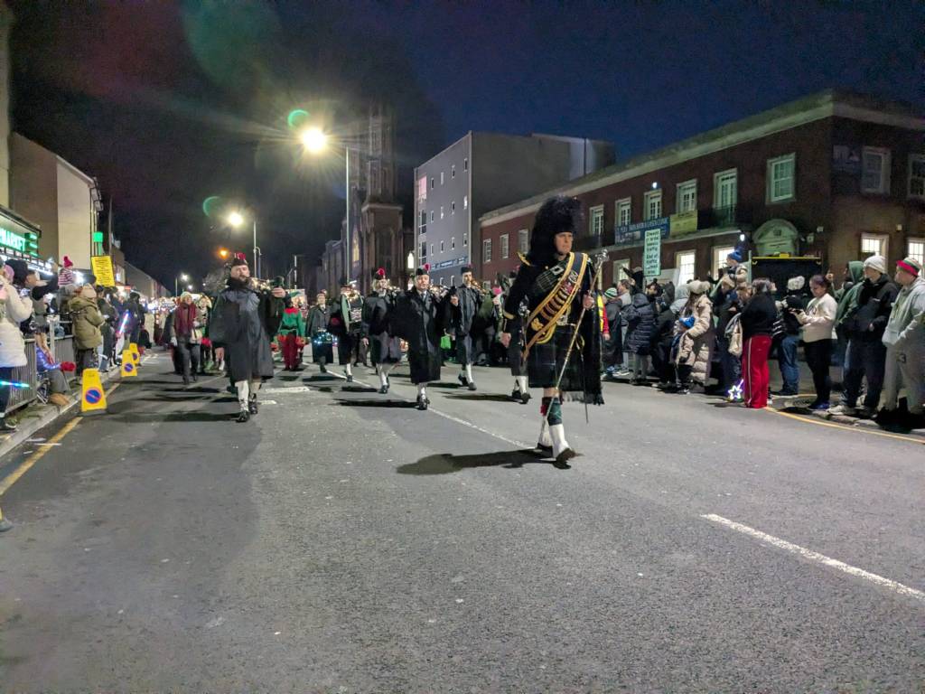 Parade participants in kilts and feathered hats playing bagpipes during festive procession.