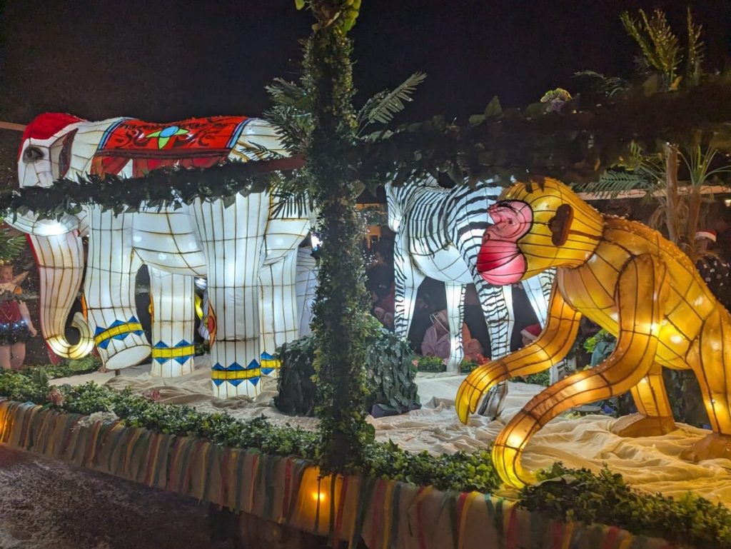Parade float with illuminated elephant, zebra, and monkey lanterns surrounded by greenery and ribbons.