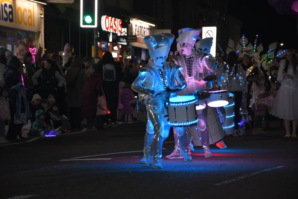 Drummers in illuminated metallic outfits performing in front of cheering crowd during parade.