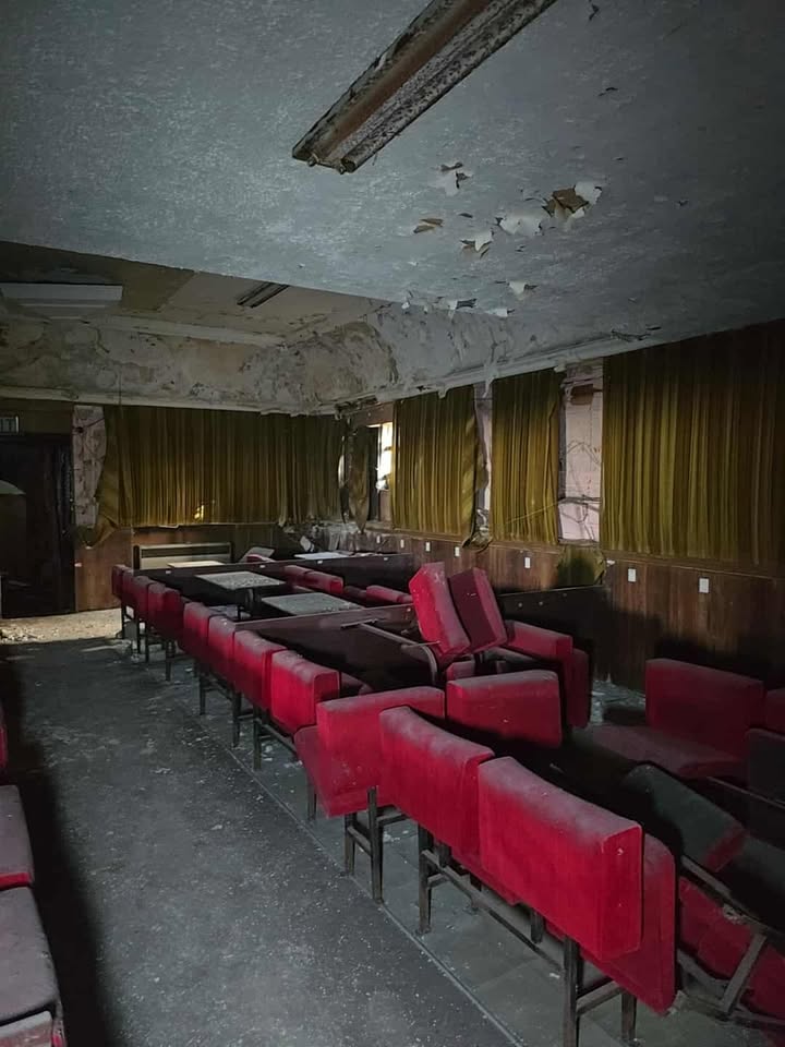 Abandoned lounge with red chairs, damaged ceiling, and debris-covered floor.