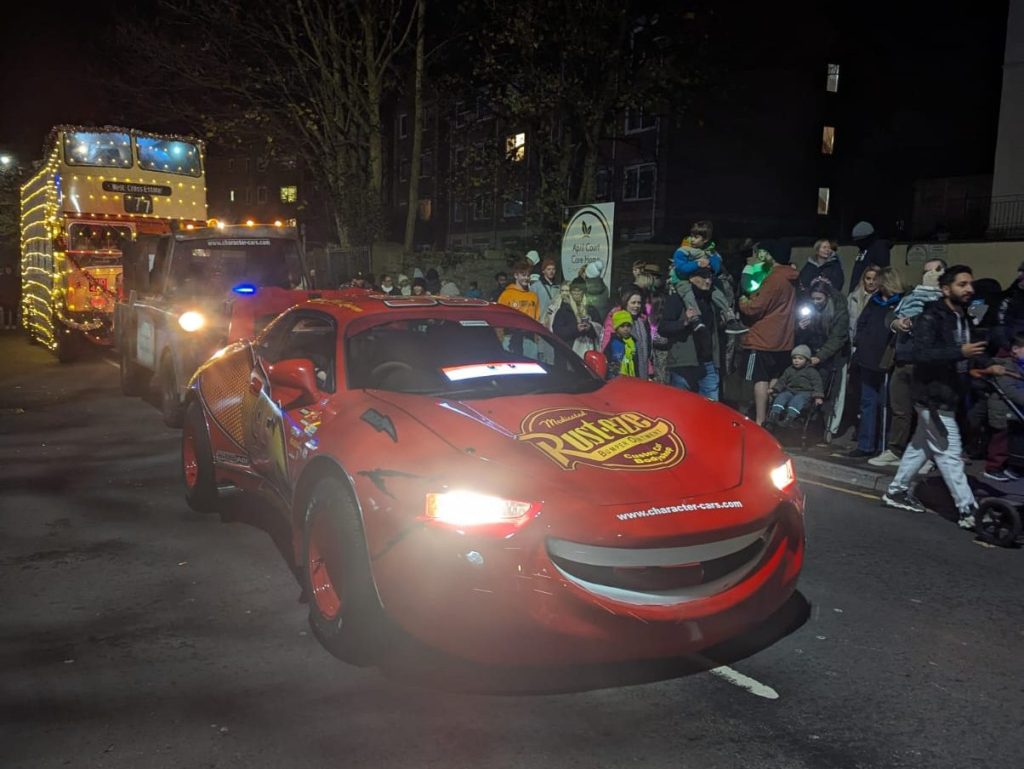 Red Lightning McQueen-style car and illuminated double-decker bus in festive parade.
