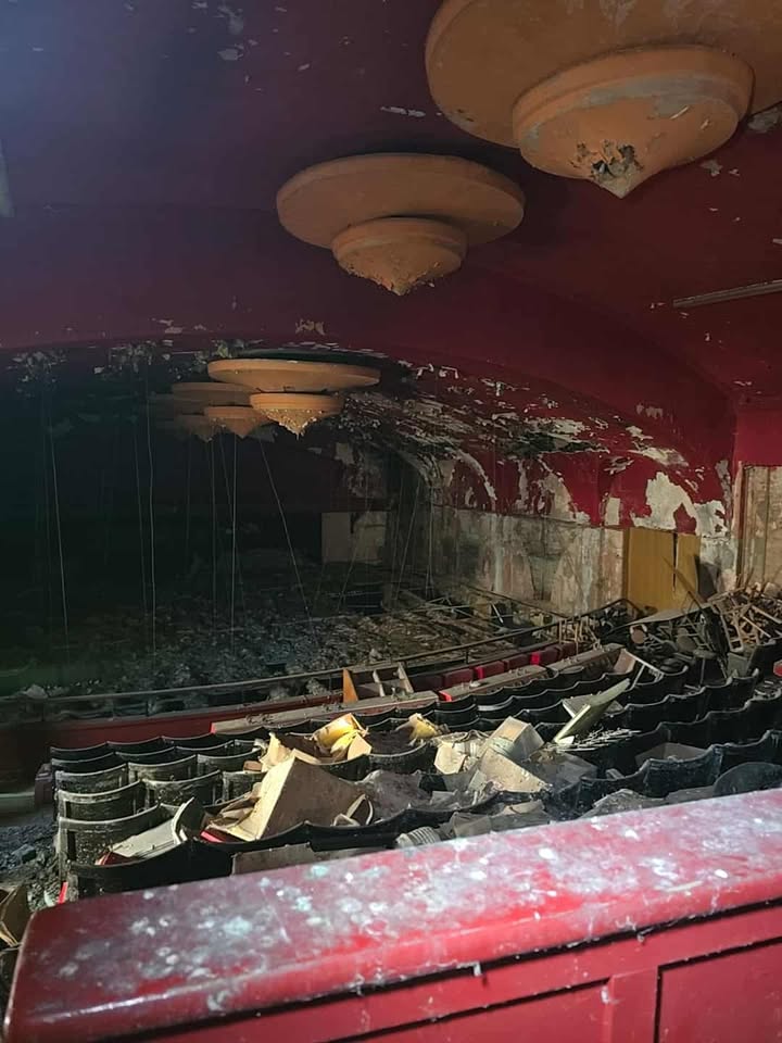 Abandoned theatre with broken seats, red ceiling, and debris covering the stage area.