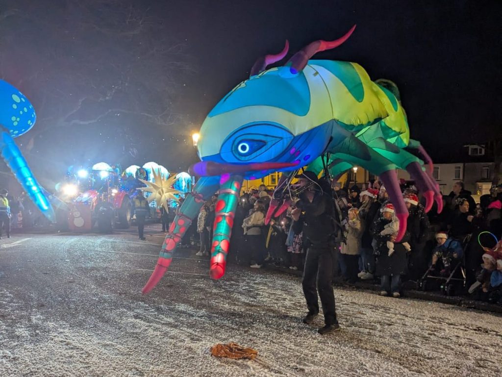 Large illuminated insect puppet with glowing eyes and antennae paraded through snow-covered street.