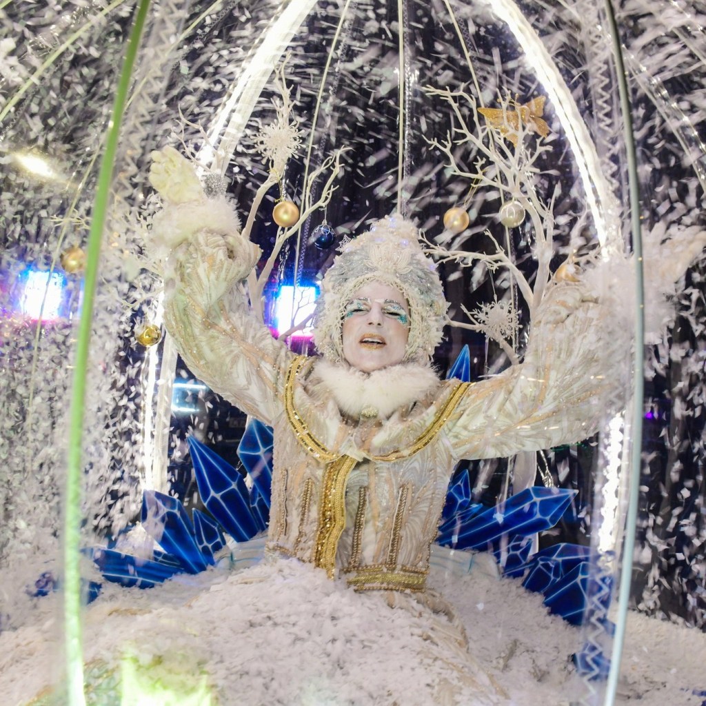 Performer dressed as Snow Queen inside illuminated snow globe with wintry decorations.