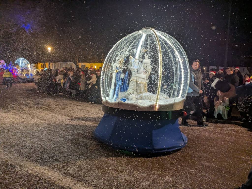 Illuminated snow globe with castle and snow-covered trees, surrounded by spectators.