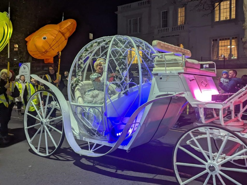 White illuminated Cinderella-style carriage with costumed performer inside, surrounded by fish lanterns.