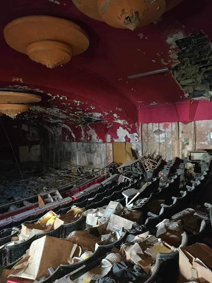 Abandoned theatre with broken seats, red ceiling, and debris covering the stage area.