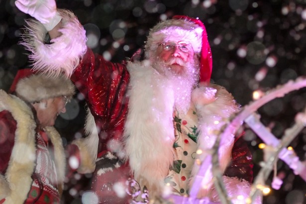 Santa Claus waving in snowfall, surrounded by lights and holiday decorations.