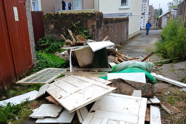 Pile of dumped household waste in an alleyway, including white doors, a bathtub, carpet pieces and timber, with a person walking away in the distance.
