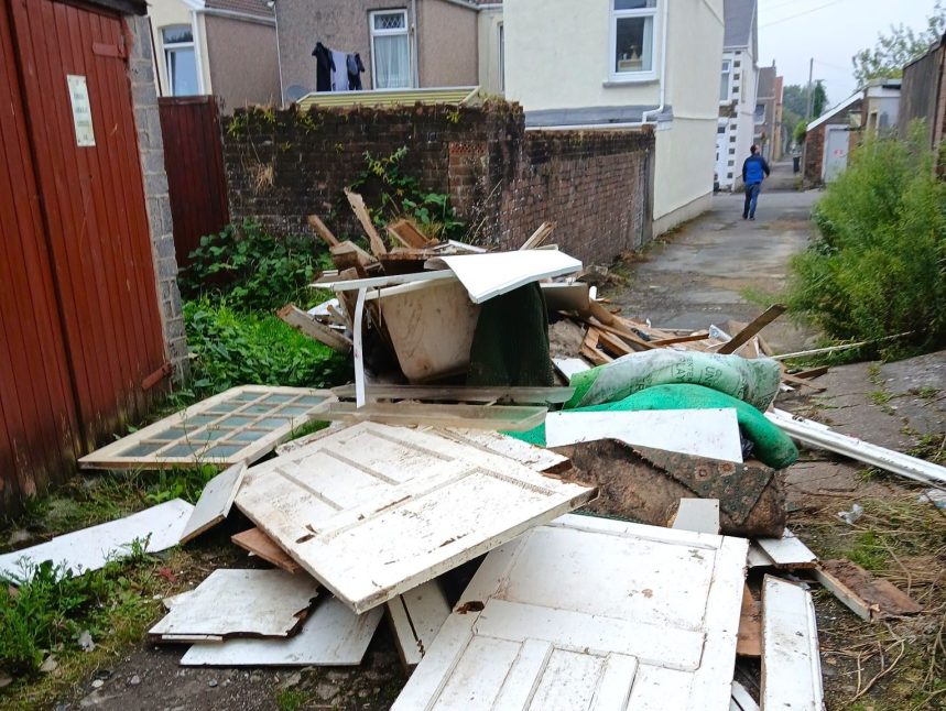 Pile of dumped household waste in an alleyway, including white doors, a bathtub, carpet pieces and timber, with a person walking away in the distance.