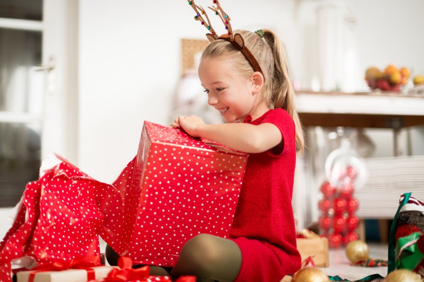 Child in red dress and reindeer antlers opens a large red gift box with white stars, surrounded by Christmas decorations and presents.