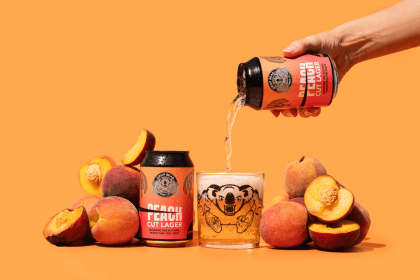 Hand pouring Drop Bear Beer Co.’s Peach Cut Lager into a branded glass, surrounded by fresh peaches on an orange background. The alcohol-free beer has a foamy head and a bear illustration on the glass.