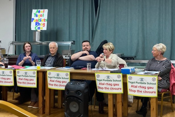 Close‑up of Pontyates school governors at a public meeting, sitting at a table with protest banners in the background.