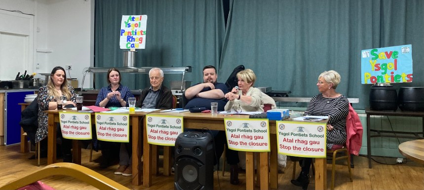 Close‑up of Pontyates school governors at a public meeting, sitting at a table with protest banners in the background.