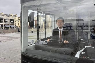 Tom Giffard MS sitting in the driver’s seat of a green electric double‑decker bus.