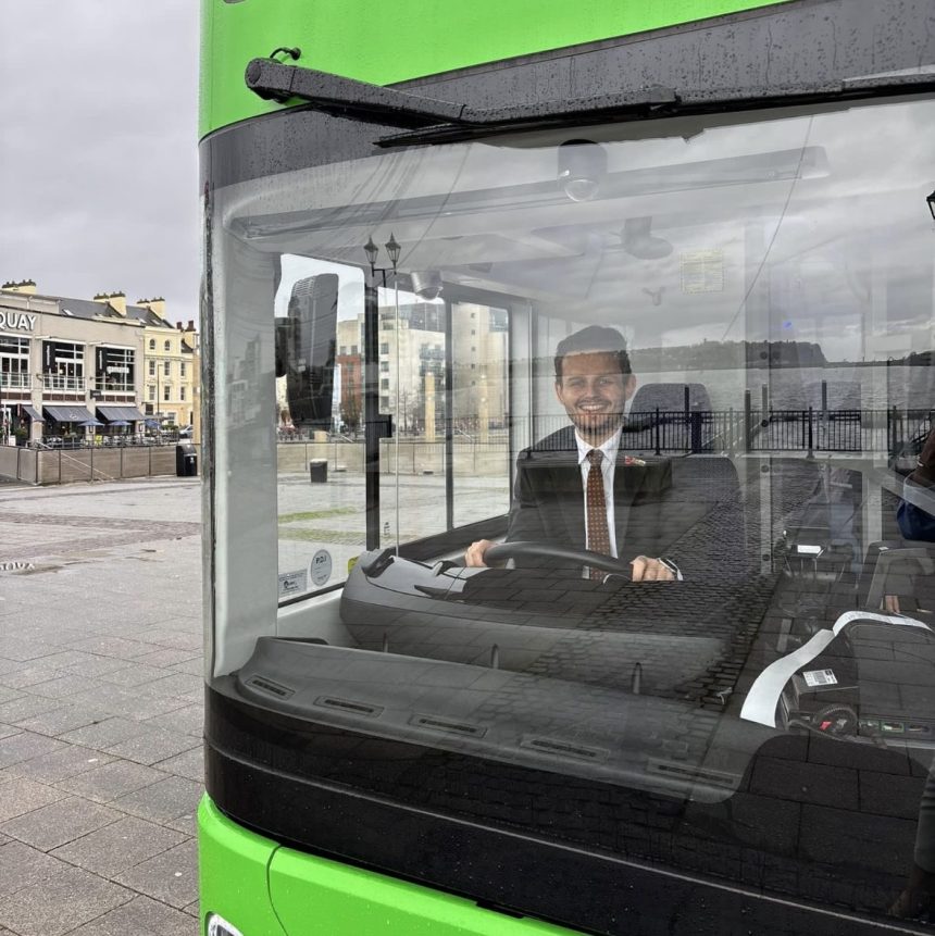 Tom Giffard MS sitting in the driver’s seat of a green electric double‑decker bus.