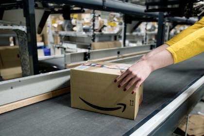 Amazon‑branded box on a rubber conveyor at the Swansea Distribution Centre, with a worker’s hand in a yellow sleeve guiding it, warehouse equipment visible behind.