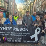 Group of people holding a White Ribbon banner and posters on a Swansea street, including a police officer and campaigners promoting domestic abuse awareness.