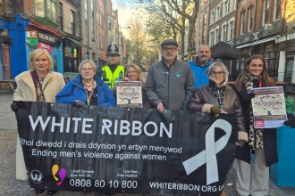 Group of people holding a White Ribbon banner and posters on a Swansea street, including a police officer and campaigners promoting domestic abuse awareness.