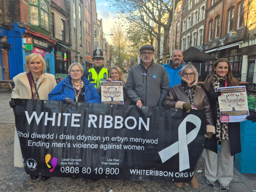 Group of people holding a White Ribbon banner and posters on a Swansea street, including a police officer and campaigners promoting domestic abuse awareness.