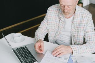 an elderly man looking at the documents on the table near his laptop