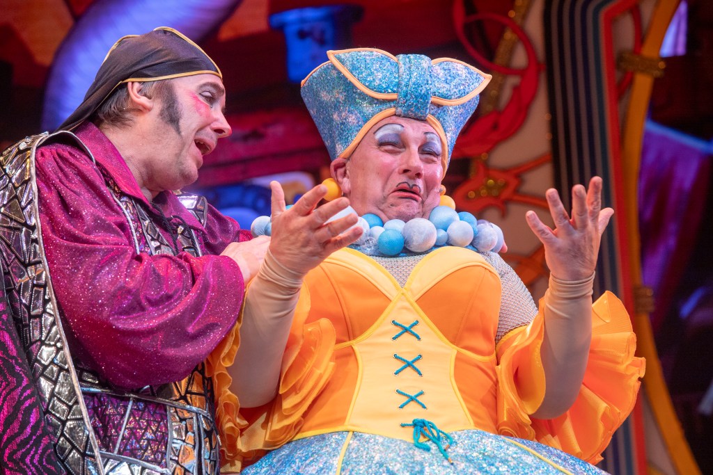 Two performers in glittering panto costumes on stage, including Kev Johns in yellow and blue headpiece.