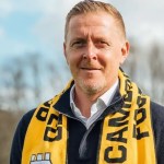 Gary Monk standing outdoors wearing a Cambridge United scarf and dark zip-up sweater, with trees in the background — image credited to Cambridge United FC.