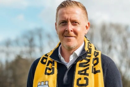 Gary Monk standing outdoors wearing a Cambridge United scarf and dark zip-up sweater, with trees in the background — image credited to Cambridge United FC.