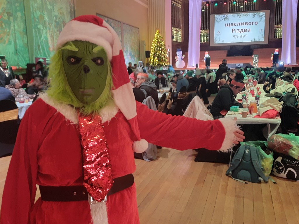 Person in Grinch costume at festive community event in Brangwyn Hall with Christmas tree and decorations.