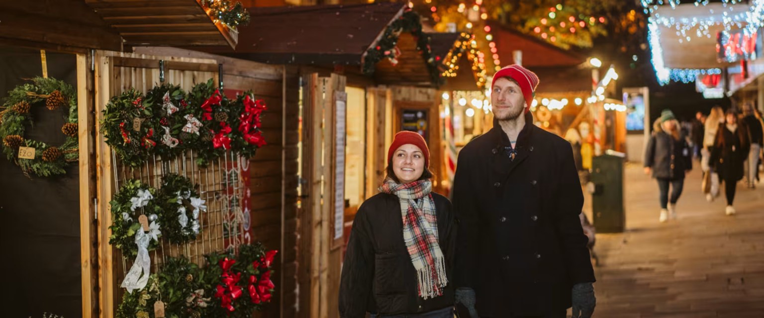 Two people walk past wreath stalls at Swansea Christmas Market, surrounded by lights, garlands and festive decorations.