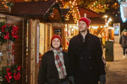 Two people walk past wreath stalls at Swansea Christmas Market, surrounded by lights, garlands and festive decorations.
