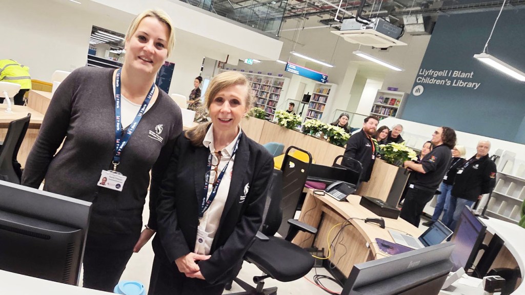 Two staff members smiling in the Children’s Library section at Y Storfa, with books and workstations behind.