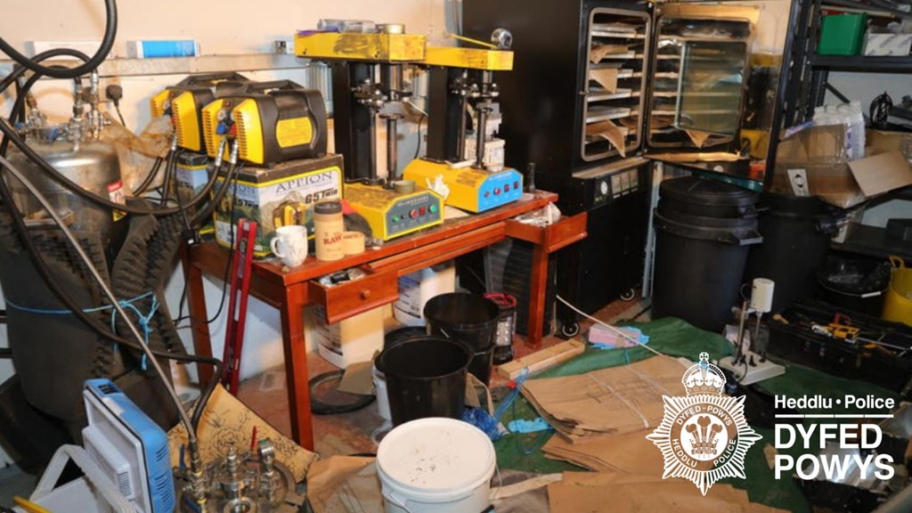Cluttered room with industrial equipment including ovens, press and containers — cannabis lab evidence.