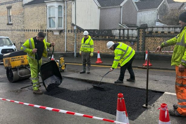 Four workers in high-vis gear repair a road section with asphalt and a Mecalac roller near Brynmill.