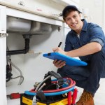 Plumber kneeling beside open kitchen sink cabinet with tools and clipboard, inspecting pipework.
