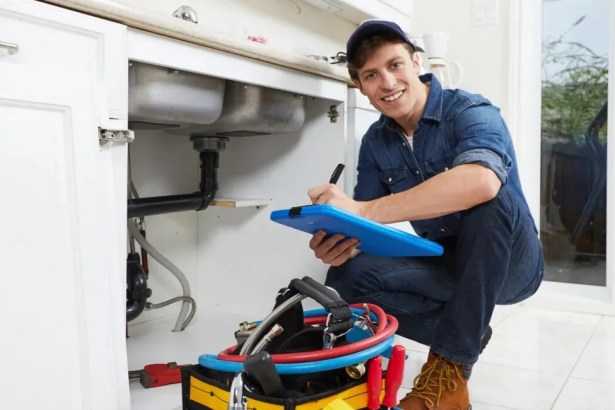 Plumber kneeling beside open kitchen sink cabinet with tools and clipboard, inspecting pipework.