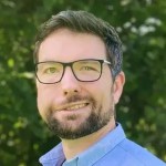 Portrait of Aaron Jones, a bearded man in glasses and a light blue shirt, photographed outdoors in natural light.