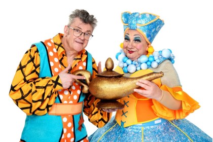 Joe Pasquale and Kev Johns in colourful pantomime costumes holding a genie lamp, promoting Aladdin at Swansea Grand Theatre. Credit: Swansea Grand / Imagine Theatre