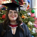 Ayla Jones smiling in graduation cap and gown beside a decorated Christmas tree, celebrating her nursing degree from Swansea University.