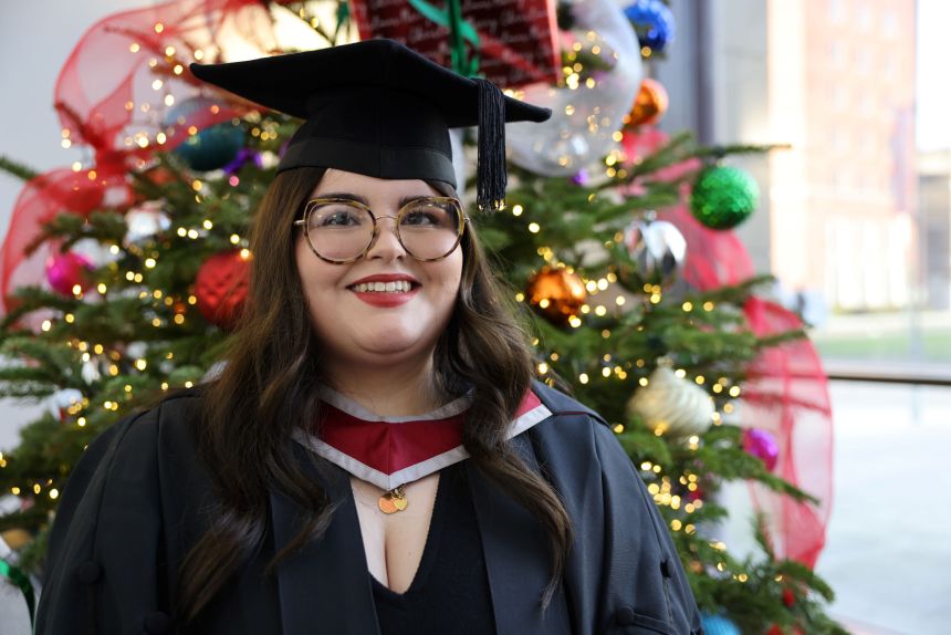 Ayla Jones smiling in graduation cap and gown beside a decorated Christmas tree, celebrating her nursing degree from Swansea University.