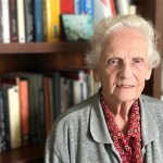 Elderly woman with white hair standing in front of a bookshelf filled with books and model ships, wearing a grey cardigan and red patterned shirt, in a study setting.