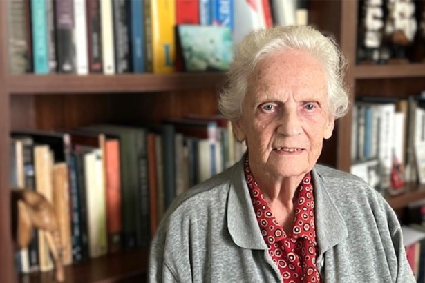 Elderly woman with white hair standing in front of a bookshelf filled with books and model ships, wearing a grey cardigan and red patterned shirt, in a study setting.