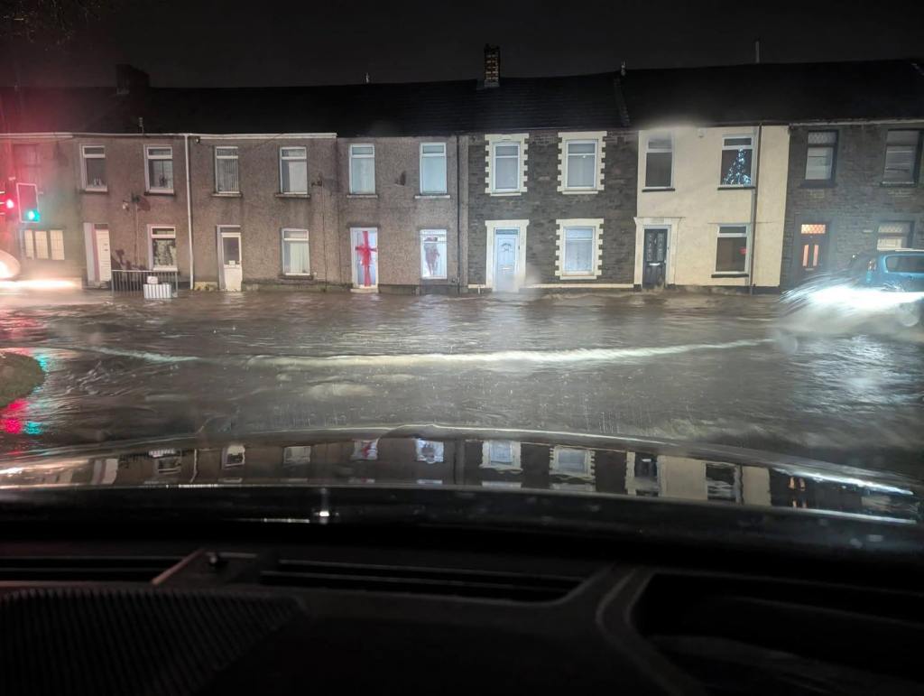 Nighttime street scene in Briton Ferry with floodwater covering the road as a car drives through, creating splashes.