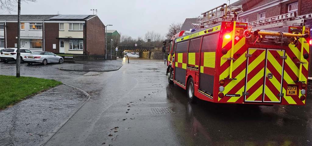Fire engine with flashing lights parked near a flooded railway bridge in Briton Ferry.