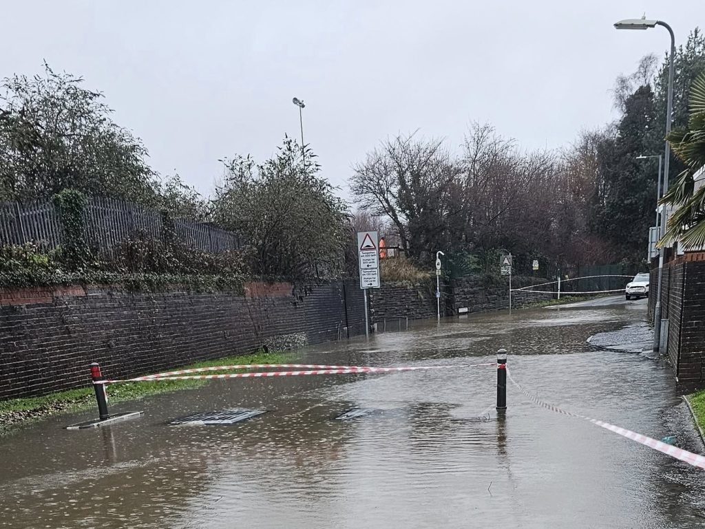 Flooded urban street in Briton Ferry with red and white caution tape blocking access.